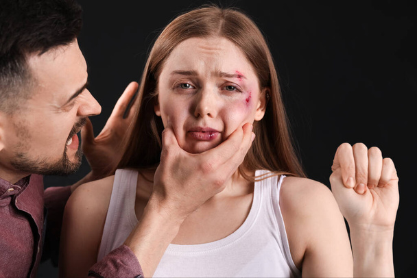 Mujer joven con moretones con su marido enojado mostrando puño sobre fondo oscuro. Concepto de violencia doméstica - Foto, Imagen