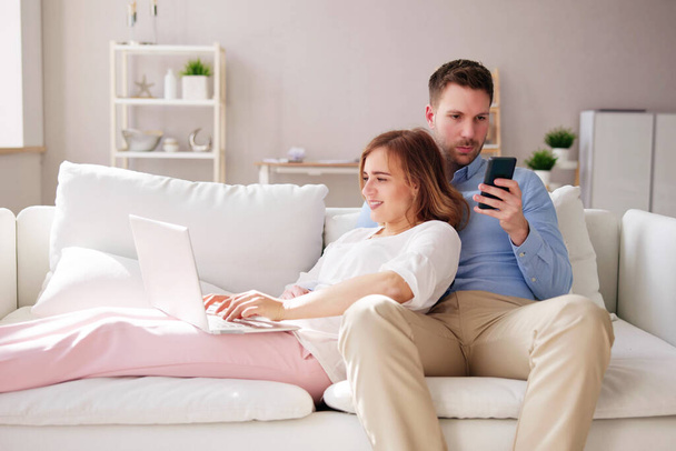 Para rodzinna korzystająca z laptopa i telefonu na kanapie - Zdjęcie, obraz