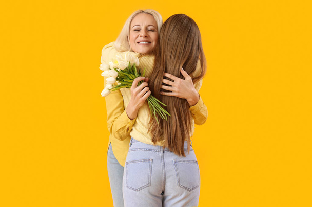 Rijpe vrouw met tulpen die haar dochter omhelst op oranje achtergrond. Moederdag - Foto, afbeelding