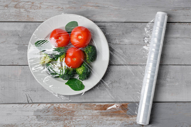 Plate with vegetables wrapped with stretch wrap on grey wooden background - Photo, Image