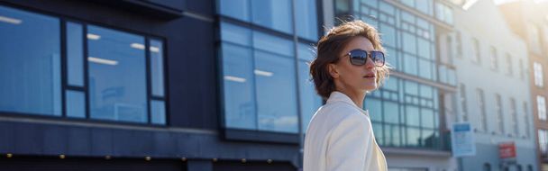 Beautiful woman in trendy outfits crosses road against backdrop of city buildings and looking away - Фото, изображение