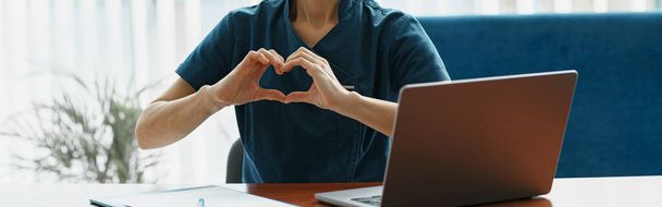 Hymyilevä lääkäri työskentelee kannettavan ja osoittaa sydämen kädet lääketieteellisessä toimistossa. Laadukas kuva - Valokuva, kuva