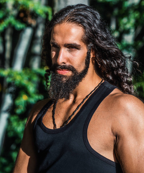 Handsome bearded man with long dark hair. Sexy bearded model man. Man fashion and style concept. Portrait of handsome young strong man with long dark hair looking away while standing at forest - Foto, afbeelding