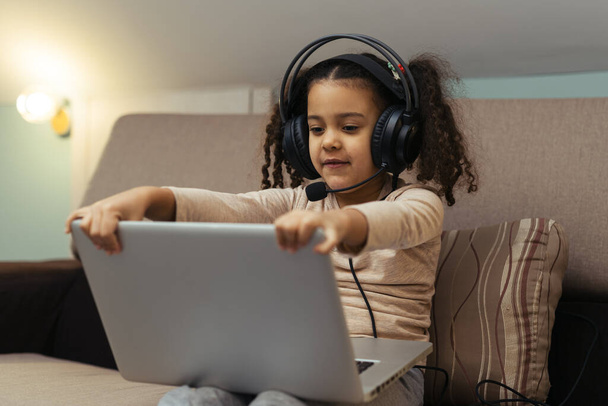 The happy little girl in headphones sitting at the sofa with a laptop - Fotoğraf, Görsel