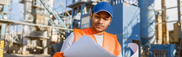Multiracial experienced worker wearing construction vest and gloves examining blueprint when standing in plant for construction industry production - Photo, Image