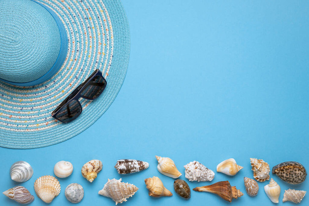 Summer and vacation flat lay with a beauty blue striped woman straw hat, sun glasses and various seashells at the lower edge of the picture on blue background. - Fotografie, Obrázek