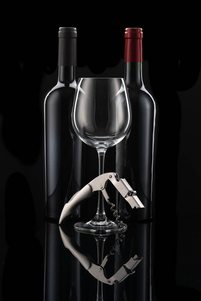 Two wine bottles next to each other, a wine glass and wine cutlery in front of a black background - Фото, изображение