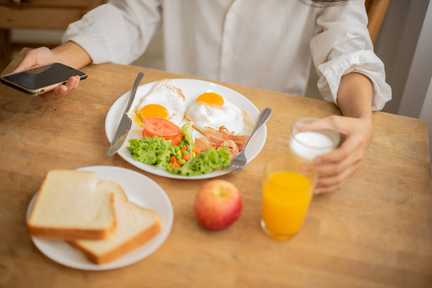 Unknown person holding mobile phone and glass of milk and breakfast on table full of fresh fruit and bread. - 写真・画像
