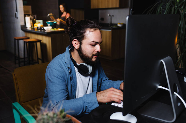 Fiatal latin férfi dolgozik és használja a számítógépet, míg a barátnő főz a háttérben otthon éjszaka Mexikóban Latin-Amerikában, spanyol emberek - Fotó, kép