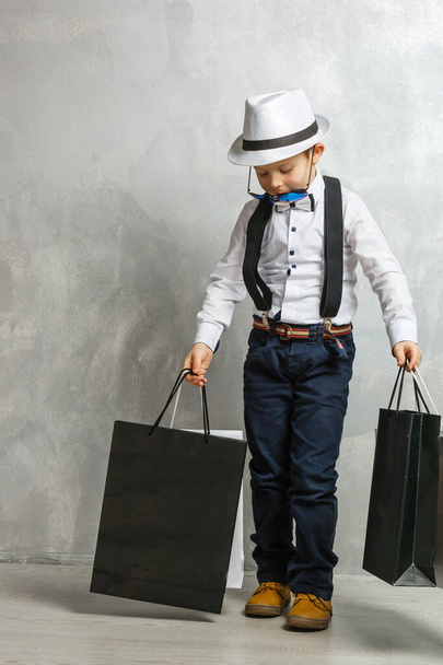 bájos kisfiú megy bevásárló show táskák vásárolni alkalmi ruhák szürke színű háttér. - Fotó, kép