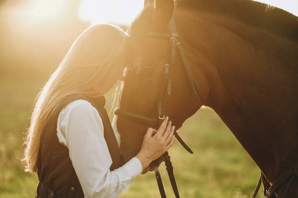 Young beautiful female jockey strokes and hugs the horse's head and prepares for the competition. Jumping training in the meadow in summer evening. - Fotoğraf, Görsel