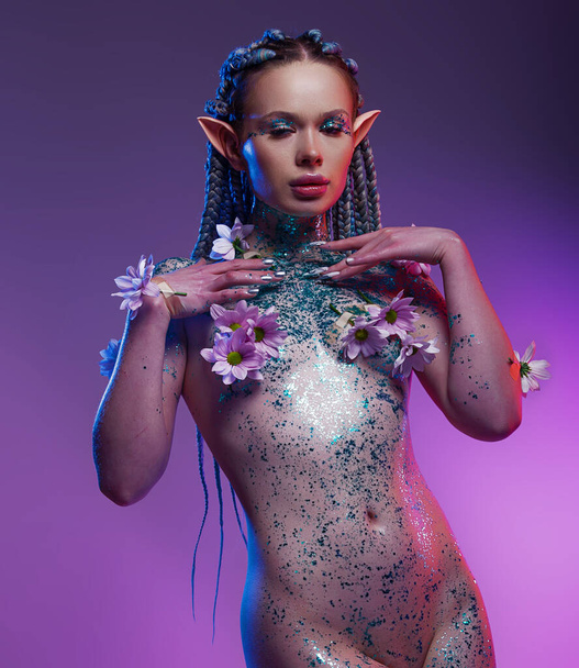 A charming female elf. One with nature and organically purity. The nude model her body covered flowers and sparkle glitter on a violet background - Photo, Image