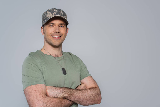 ευτυχισμένος Αμερικανός στρατιώτης με t-shirt και στρατιωτικό καπέλο στέκεται με διπλωμένα χέρια κατά τη διάρκεια της ημέρας μνήμης απομονώνονται σε γκρι - Φωτογραφία, εικόνα