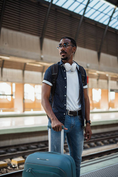 Hombre africano sosteniendo una maleta azul en una estación de tren mientras espera a que llegue el tren - Foto, imagen