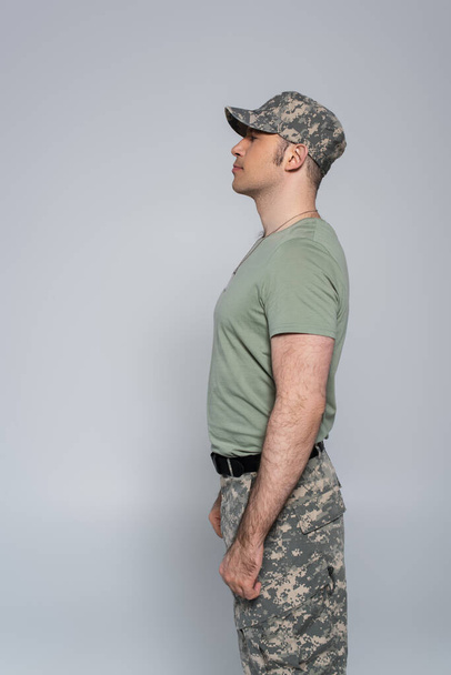 boční pohled vojáka v tričku a vojenské čepici stojící během památného dne izolovaný na šedi  - Fotografie, Obrázek