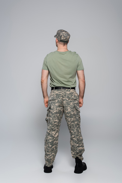 πίσω όψη του στρατιώτη με στρατιωτική στολή και καπέλο όρθιο σε γκρι  - Φωτογραφία, εικόνα