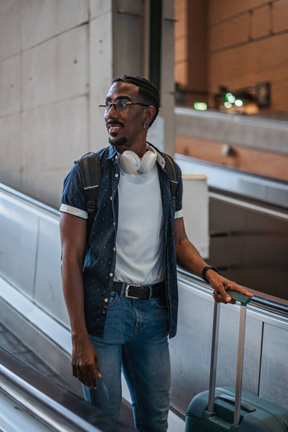 Portrait of black young man on a ramp escalator in a train station. He is holding his suitcase - 写真・画像
