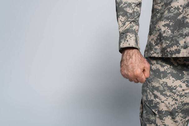 przycięty widok patriotycznego żołnierza w mundurze wojskowym stojącego z zaciśniętą pięścią na szarym tle  - Zdjęcie, obraz
