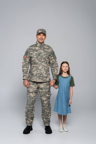 full length of happy army soldier in military uniform standing with daughter during memorial day on grey - Fotoğraf, Görsel