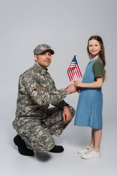 cheerful preteen girl giving flag of United States to happy father in military uniform during memorial day on grey  - 写真・画像