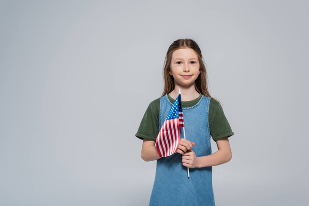 patriotique preteen fille souriant tout en tenant drapeau de l'Amérique lors de la journée commémorative isolé sur gris  - Photo, image