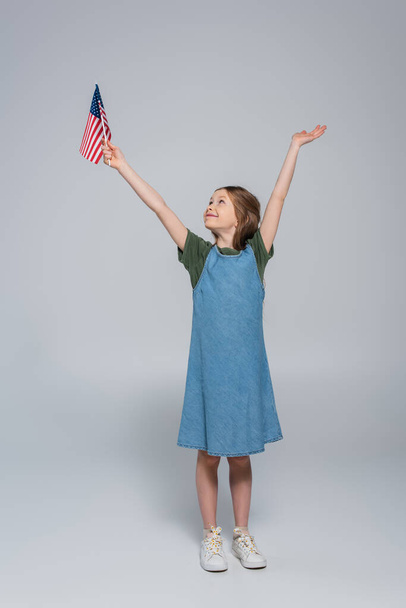 full length of patriotic preteen girl smiling while holding flag of United States of America during memorial day on grey  - Photo, image