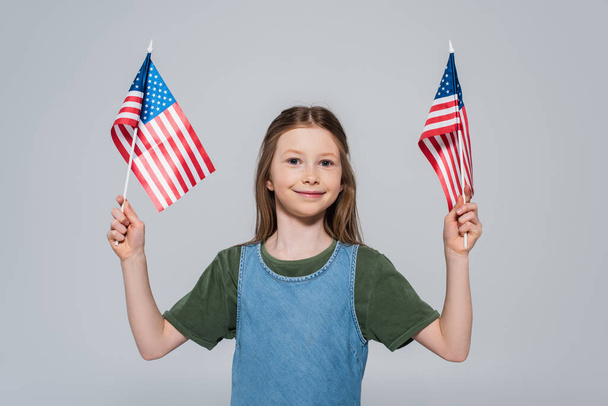 niña preadolescente patriótica sonriendo mientras sostiene banderas de América durante el día conmemorativo aislado en gris  - Foto, imagen