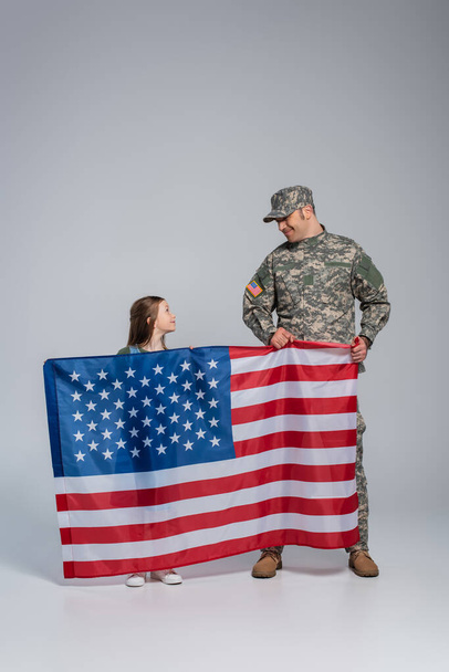 Askeri üniformalı neşeli asker, gri üzerinde mutlu kızı olan Birleşik Devletler bayrağını taşıyor.  - Fotoğraf, Görsel