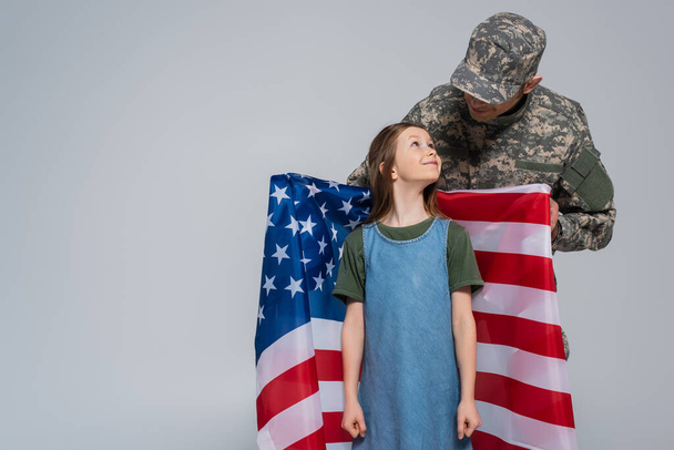 żołnierz w mundurze wojskowym trzymający flagę Stanów Zjednoczonych w pobliżu szczęśliwej córki odizolowanej na szaro  - Zdjęcie, obraz