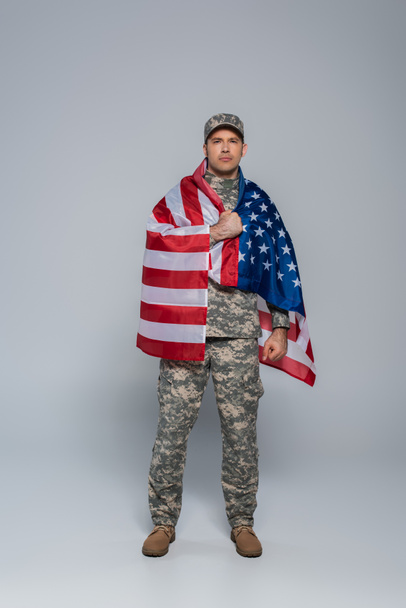 full length of patriotic army soldier in camouflage uniform wrapped in flag of United States of America during memorial day on grey  - Фото, изображение