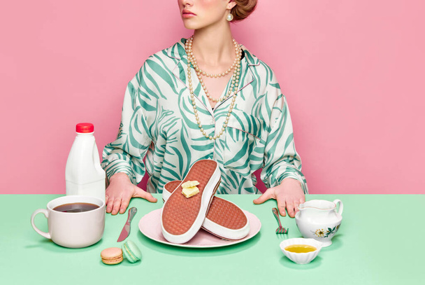 Обрізане зображення стильної дівчини, що снідає з сюрреалістичними кросівками вафлі та кава на фоні рожевої студії. Їжа поп-арт фотографія. Концепція ретро-стилю, творче бачення, уява
. - Фото, зображення