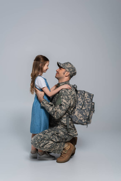 szczęśliwy żołnierz w mundurze wojskowym i plecaku przytulając wesołą córkę na jego powrót do domu na szarym tle  - Zdjęcie, obraz