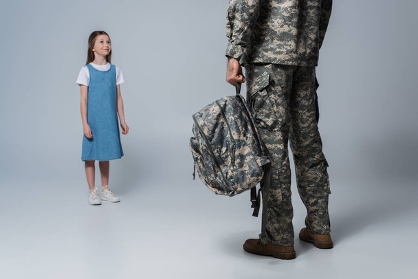 menina feliz olhando para o pai em uniforme militar segurando mochila durante o baile em fundo cinza  - Foto, Imagem