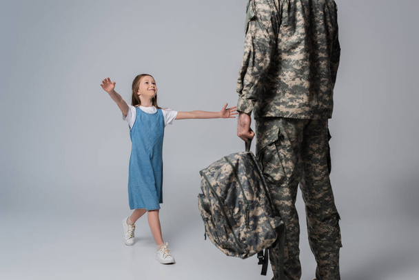 fille heureuse accueillant père en uniforme militaire avec sac à dos pendant le retour à la maison sur fond gris  - Photo, image