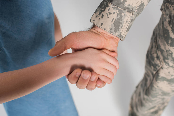 vista cortada de menina pré-adolescente segurando a mão de pai corajoso em uniforme do exército durante o dia comemorativo isolado em cinza  - Foto, Imagem