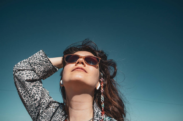 Jovem morena sorrindo mulher usando óculos de sol, jaqueta de leopardo olhando para cima contra um céu azul claro primavera. Liberdade, independência, conceito de sonho. Menina hipster mod sonhador. turista feminino. - Foto, Imagem
