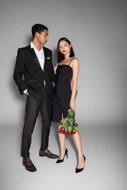 full length of african amrican man in black stylish suit embracing asian woman holding red roses on grey background - Foto, immagini
