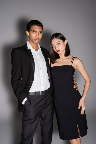 african american man in black suit and asian woman in strap dress looking at camera on grey background - Foto, Bild