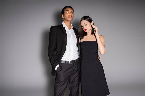 african american man in elegant suit looking at camera near asian woman in black dress on grey background - Foto, Imagem