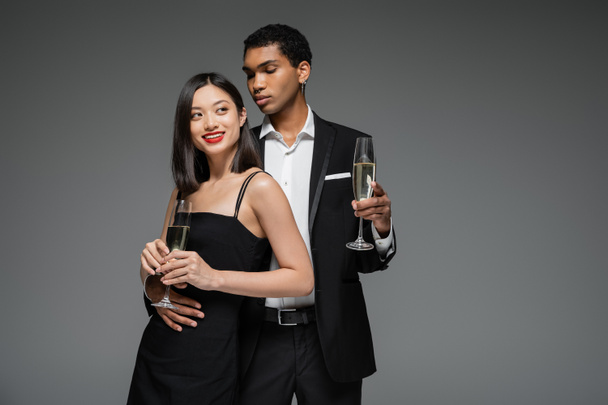 усміхнена азіатська жінка і елегантний афроамериканський чоловік тримає шампанські окуляри ізольовані на сірому
 - Фото, зображення