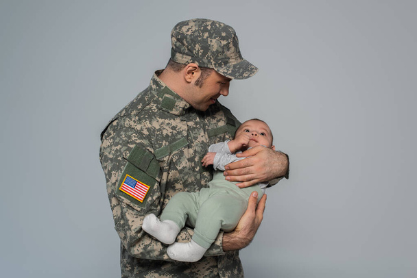 glücklicher Soldat in Uniform und Mütze, neugeborener Sohn isoliert auf grau  - Foto, Bild