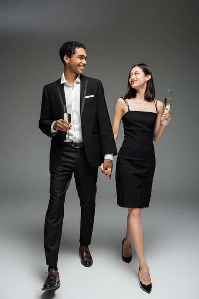 pełna długość eleganckiej wieloetnicznej pary z kieliszkami szampana trzymającej się za ręce i uśmiechającej się do siebie podczas spaceru po szarym tle - Zdjęcie, obraz