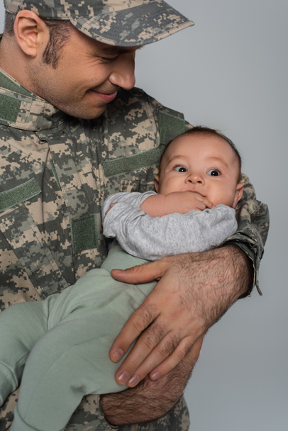 szczęśliwy żołnierz w mundurze wojskowym i czapce trzymając nowonarodzonego syna odizolowanego na szaro  - Zdjęcie, obraz