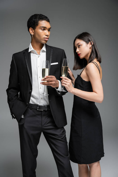elegant african american man in suit and asian woman in black dress posing with champagne glasses isolated on grey - Valokuva, kuva