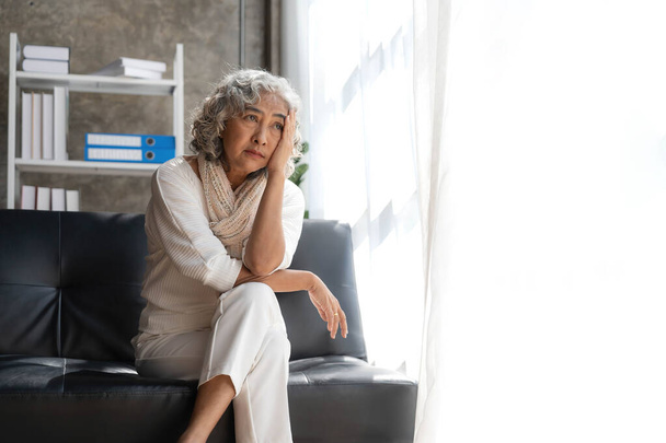 Depressed Asian senior woman sit on sofa looking away by the window. Loneliness elderly female living alone at home with sadness face. Retirement mental health care and illness recovery concept.. - Photo, Image