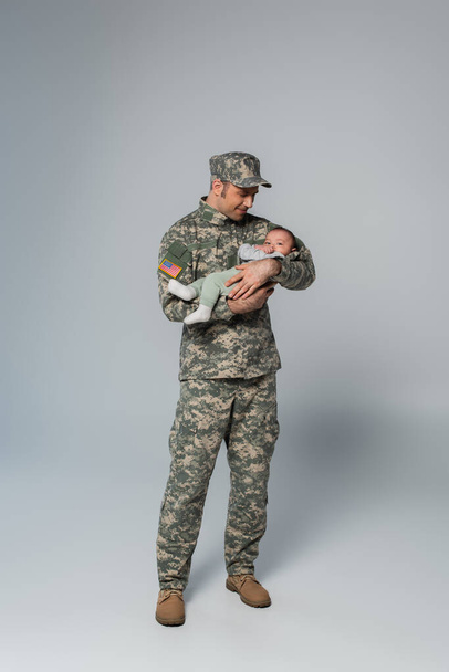 πλήρες μήκος του χαρούμενου στρατιώτη με στολή και καπέλο κρατώντας νεογέννητο γιο σε γκρι  - Φωτογραφία, εικόνα