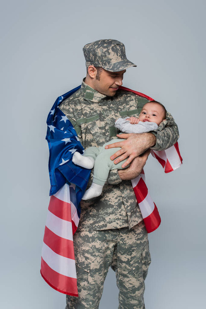 patriotyczny żołnierz w mundurze i czapce stojący z amerykańską flagą i trzymający nowonarodzonego syna odizolowanego na szaro  - Zdjęcie, obraz