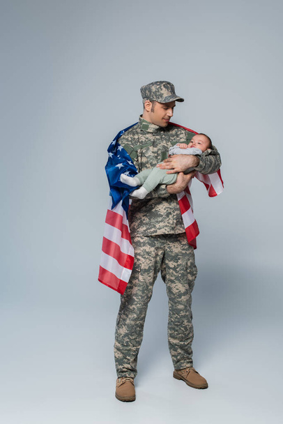 Patriotischer Soldat in Uniform und Mütze steht mit amerikanischer Flagge und hält einen kleinen Jungen in den Armen auf grau  - Foto, Bild