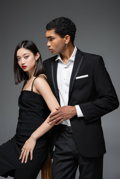 african american man in black blazer looking away near asian woman in elegant strap dress isolated on grey - Фото, зображення