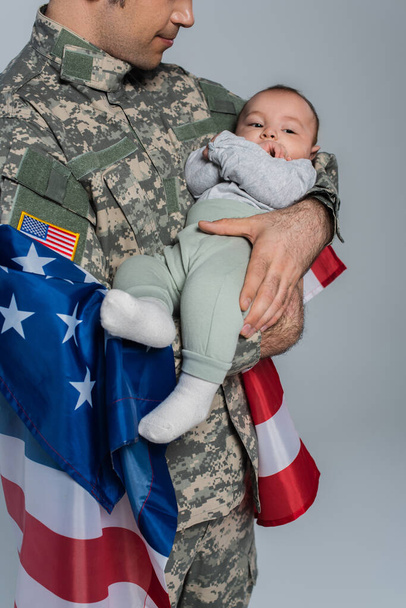 patriottische militair in uniform staat met vlag van Amerika en houdt baby jongen in de armen op grijs  - Foto, afbeelding